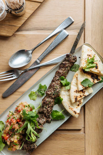 Vista superior de kebab y pan plano servido con hierbas frescas en la mesa de madera en el restaurante - foto de stock