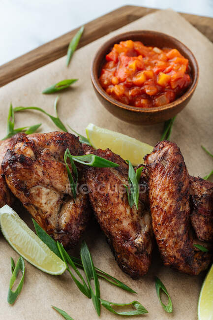 Du dessus des ailes de poulet grillées sur une planche de bois avec des tranches d'herbes de lime et de sauce rouge — Photo de stock