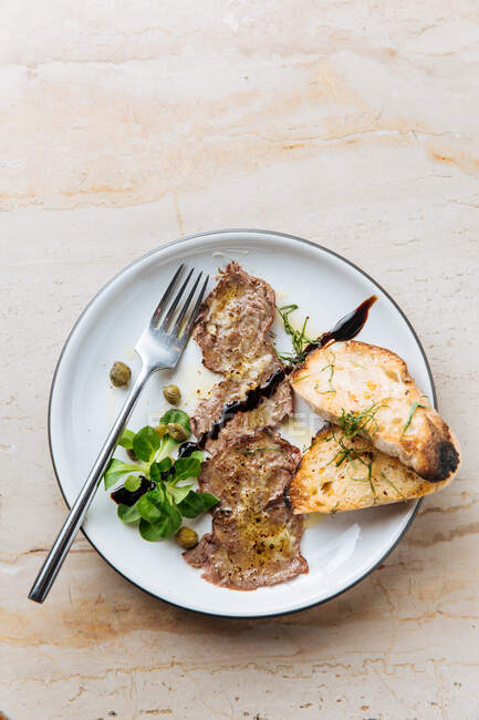 Vista dall'alto di gustosi pezzi di carne fritti su piatto bianco con pane arrosto decorato con salsa verde e forchetta metallica nel ristorante — Foto stock