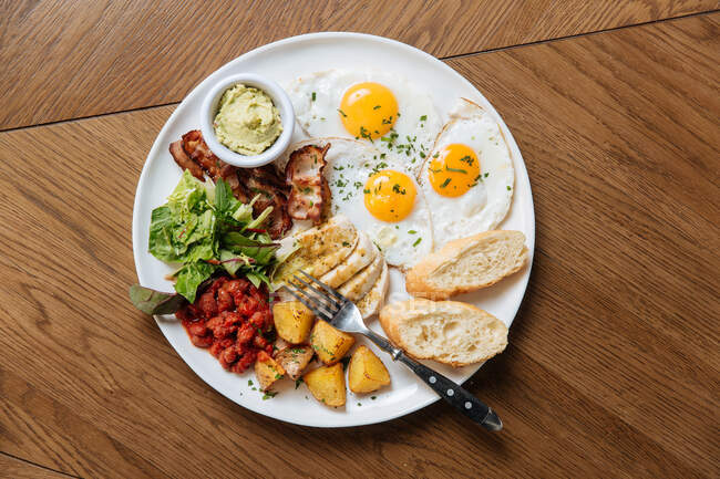 Вид сверху подаваемого английского завтрака с жареными яйцами и беконом с тостами и овощами на деревянном столе — стоковое фото
