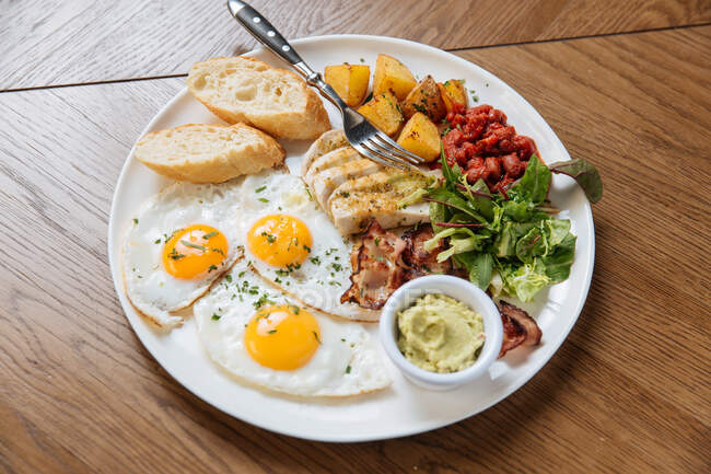 Вид сверху подаваемого английского завтрака с жареными яйцами и беконом с тостами и овощами на деревянном столе — стоковое фото