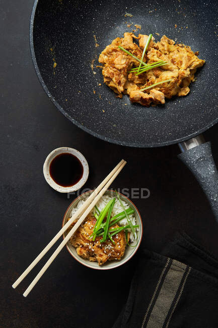 Vista superior de pollo frito en la sartén y el tazón con plato tradicional japonés Oyakodon servido con arroz hervido hierbas frescas salsa de soja y palillos - foto de stock
