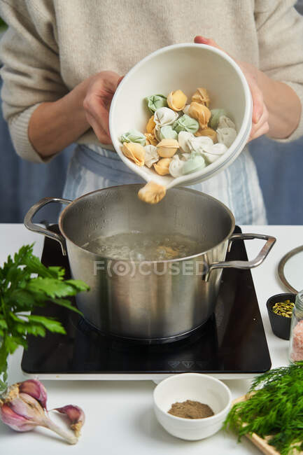 Faceless woman in apron pouring dumplings made from dough with carrots and spinach into pan with boiling water on table wish fresh herbs and species — Stock Photo