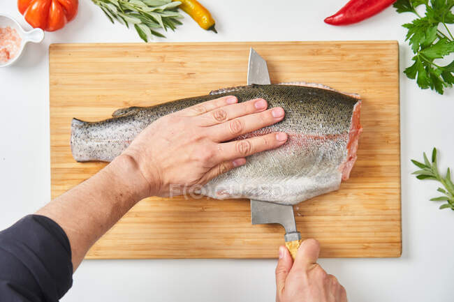 Vista superior das mãos de colheita do chef irreconhecível cortando peixe cru ao meio com faca na tábua de madeira na mesa branca com ervas frescas pimentas e espécies — Fotografia de Stock