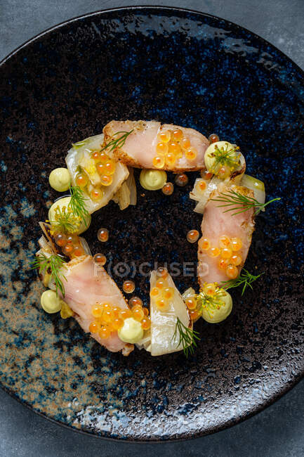 Top view of fillet of fish and red caviar decorated with cream sauce leading out in circle on stylish black plate — Stock Photo
