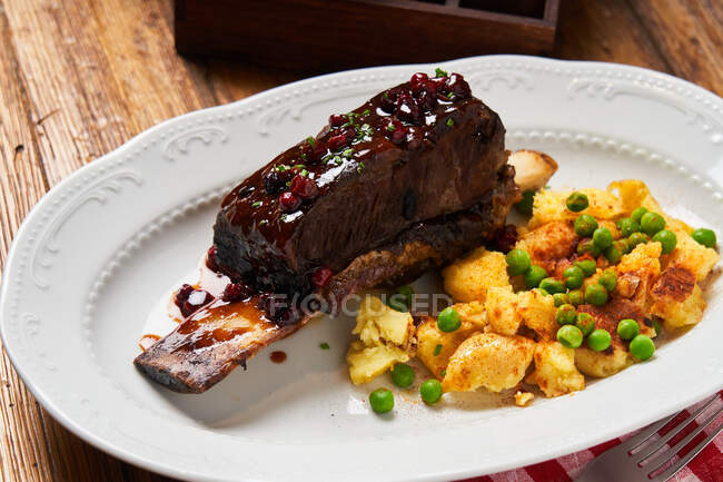From above of roasted steak on bone drizzled with berry sauce and baked golden potatoes with young green peas on white plate — Stock Photo
