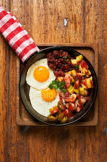 Vue du dessus des œufs frits avec des morceaux de viande et légumes dans une casserole sur une table en bois — Photo de stock