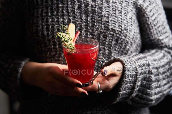 Femme anonyme en pull décontracté tenant verre avec poinçon rouge décoré de romarin et tranches de gingembre — Photo de stock