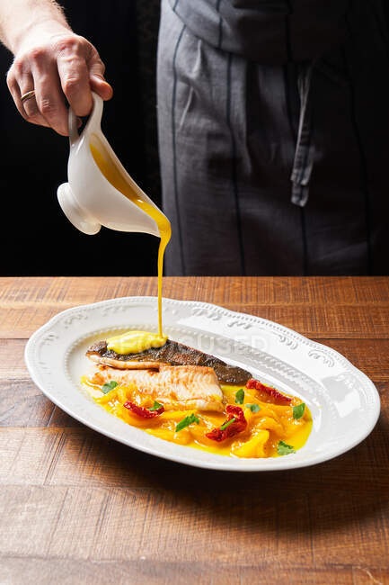 Chef masculino sin rostro en delantal usando un pequeño cruet de cerámica mientras se vierte con salsa gruesa amarilla pescado rojo guisado con verduras de colores - foto de stock