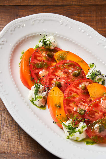 From above of fresh red slices of tomatoes and mango on white plate decorated with herbs feta cheese and pesto sauce — Stock Photo