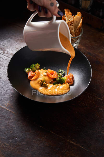 De cima da mão do chef feminino despejando prato com peixe vermelho e ervas verdes com molho de laranja em tigela preta elegante — Fotografia de Stock