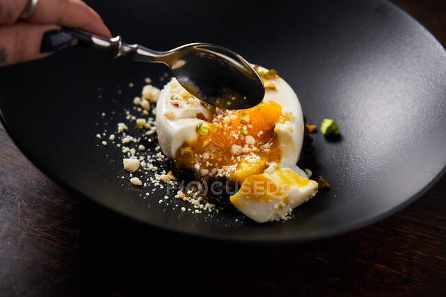 From above of crop hand with spoon of faceless woman taking piece of orange jelly glazed with white chocolate in black bowl — Stock Photo