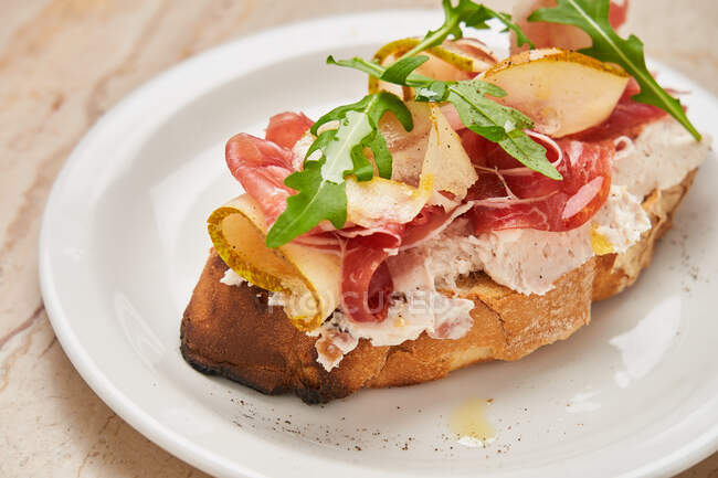 From above of ham with slices of pear on roasted bread with cream cheese decorated with arugula on white plate — Stock Photo