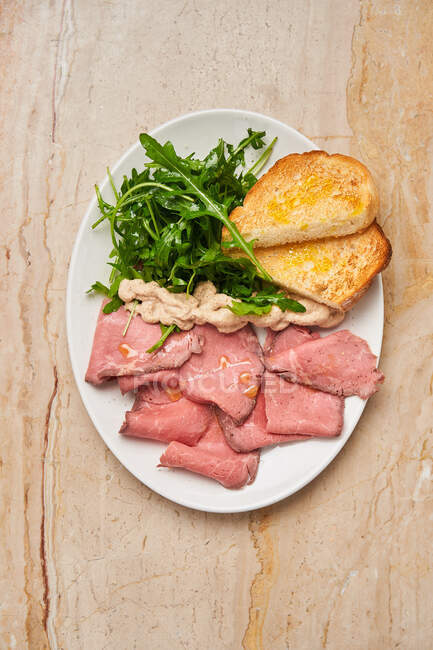 Top view of ham with slices of roasted bread and fresh arugula in white plate on wooden table — Stock Photo