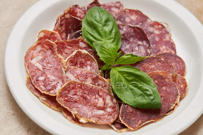 From above of white plate with slices of sausage served with green basil leaves in restaurant — Stock Photo