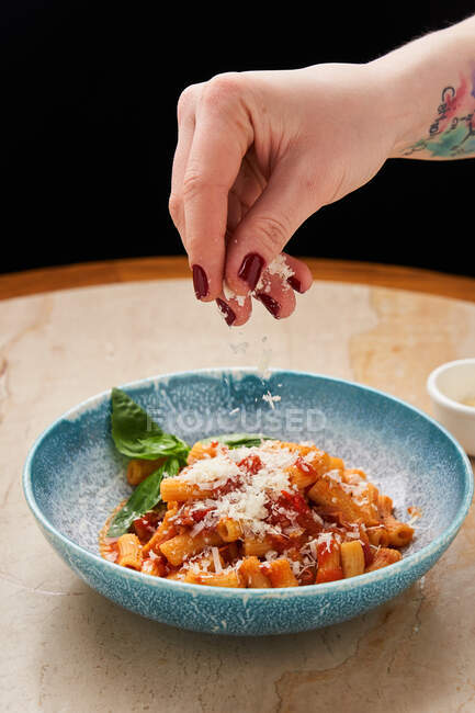 Coltivare la mano femminile cosparsa di formaggio grattugiato su tagliatelle con salsa di pomodoro e foglie di basilico in ciotola blu — Foto stock