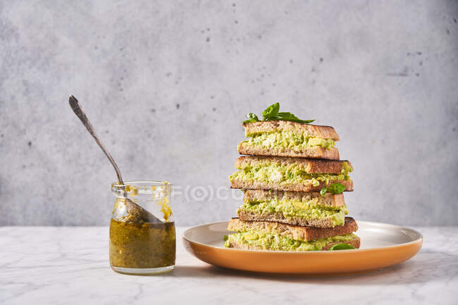 Stack of rye bread sandwiches with avocado puree served with fresh mint on table with glass jar of pesto sauce — Stock Photo