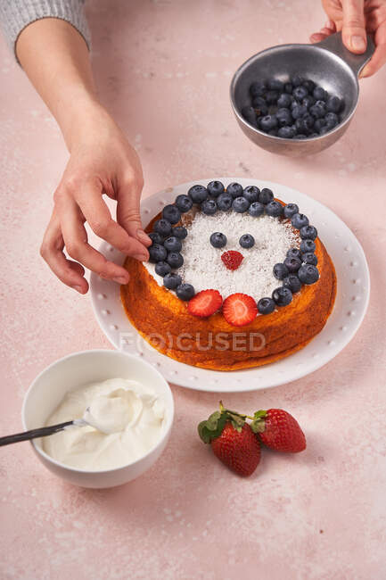 From above of unrecognizable woman decorated homemade cake with blueberry and strawberry on table with bowl of sour cream — Stock Photo