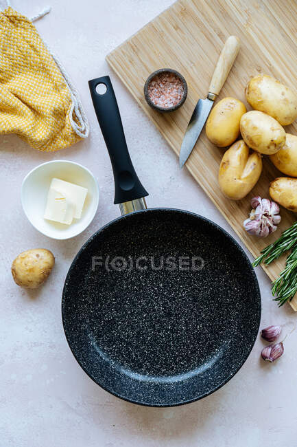 Накладні свіжі картоплі та спеції розміщені на обробній дошці біля порожньої сковороди та серветки на столі на кухні — стокове фото