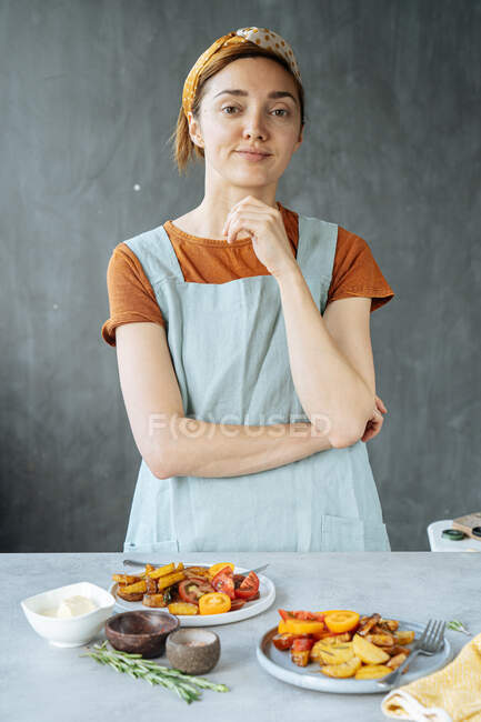 Позитивна жінка в повсякденному одязі стоїть біля столу зі смачними смаженими овочами під час приготування обіду — стокове фото