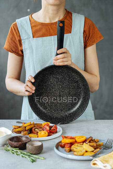 Unrecognizable female with empty frying pan standing near table with delicious roasted vegetables in kitchen — Stock Photo