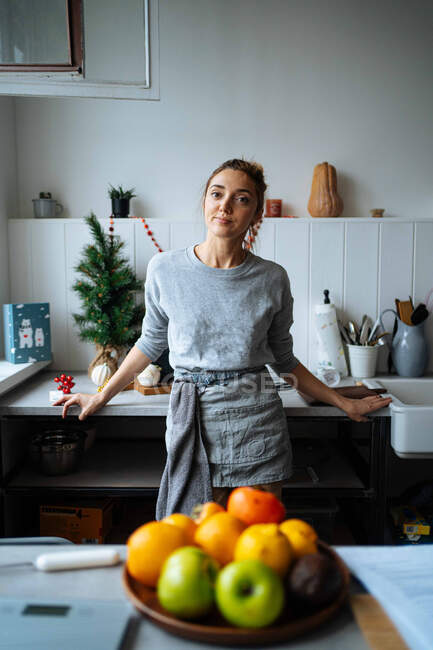 Положительная взрослая женщина в фартуке опирается на прилавок и смотрит в камеру во время приготовления пищи на кухне дома — стоковое фото