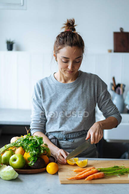 Взрослая женщина нарезает свежий лимон рядом с спелыми фруктами и овощами, стоя на кухне и готовя здоровое блюдо на обед — стоковое фото