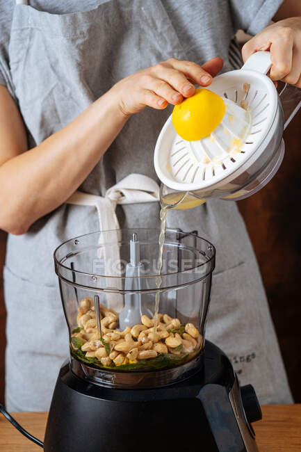 Donna irriconoscibile versando succo di limone fresco da spremiagrumi in frullatore con anacardi durante la preparazione di cibo vegan a casa — Foto stock