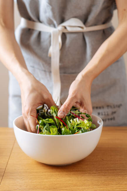 Кукурудзяна самиця в фартусі, змішуючи свіжі трави в мисці, готуючи здоровий веганський салат на столі на кухні — стокове фото
