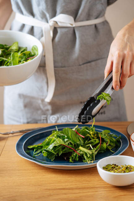 Неузнаваемая дама в фартуке, используя щипцы, чтобы разместить свежую смесь трав на тарелке во время приготовления здорового веганского салата дома — стоковое фото