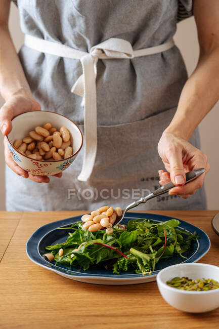 Mujer de la cosecha en delantal poniendo frijoles blancos en hierbas frescas mientras prepara ensalada vegetariana saludable para el almuerzo - foto de stock