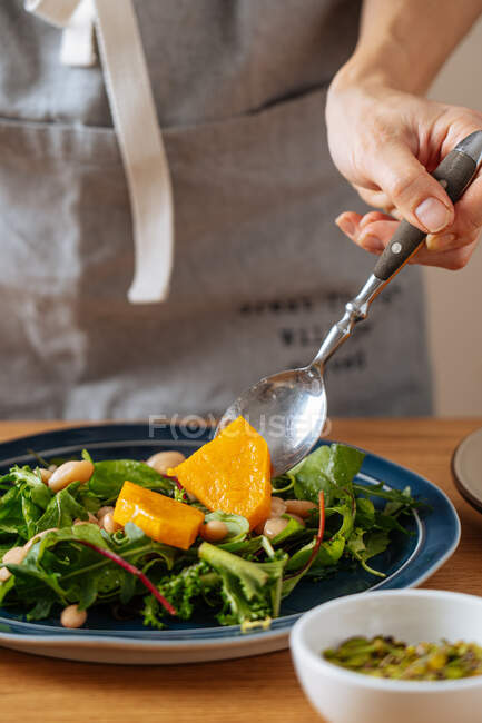 Unrecognizable person putting piece of boiled pumpkin on fresh herbs and beans while preparing healthy salad for lunch — Stock Photo