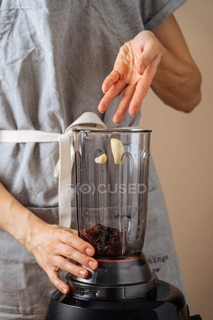 Mujer irreconocible agregando ajo fresco en la licuadora mientras cocina en la cocina en casa - foto de stock
