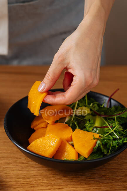 Unrecognizable person taking piece of fresh pumpkin from bowl with herbs while preparing vegan dish at home — Stock Photo