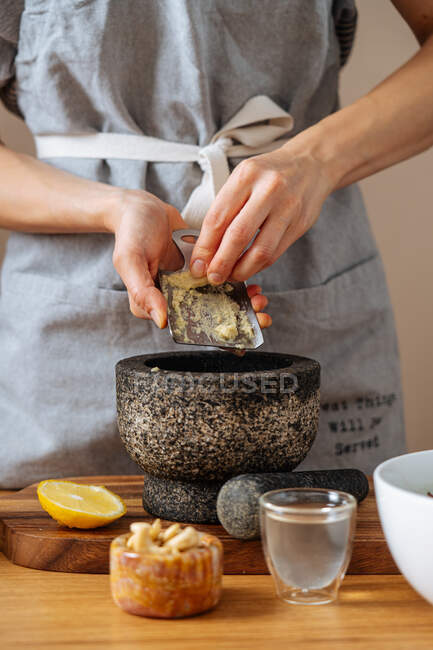 Donna irriconoscibile in grembiule macinare zenzero fresco in malta mentre si prepara piatto sano in cucina a casa — Foto stock