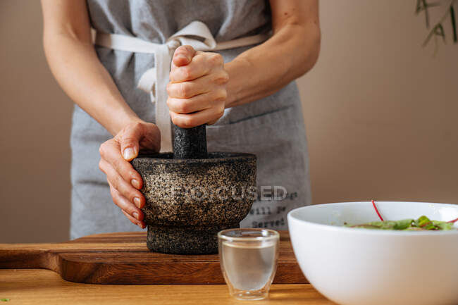 Senhora anônima em avental usando argamassa e pilão para esmagar ingredientes para molho enquanto prepara salada saudável em casa — Fotografia de Stock