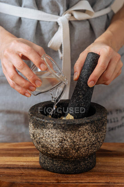 Unrecognizable person in apron spilling cup of water into mortar while preparing sauce at home — Stock Photo