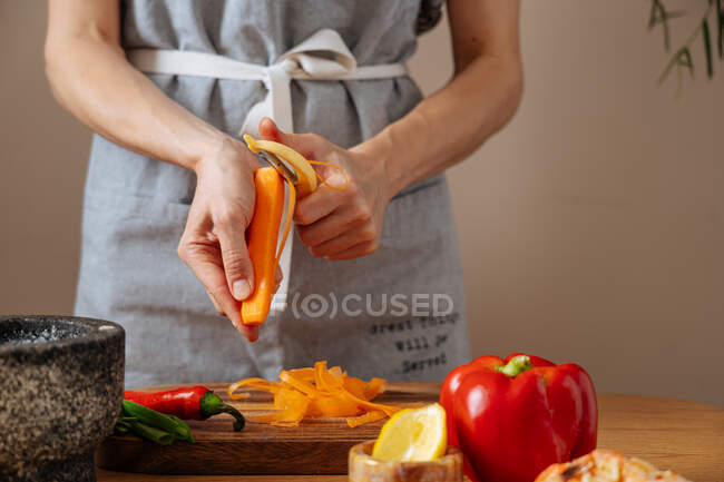 Pessoa da colheita em avental cortando cenoura fresca sobre a mesa enquanto prepara salada saudável na cozinha em casa — Fotografia de Stock