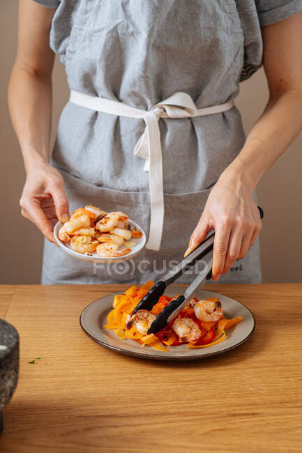 Mulher de colheita em avental usando pinças para adicionar camarões à placa com salada enquanto prepara o almoço em casa — Fotografia de Stock