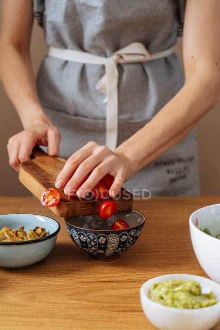 Femme méconnaissable dans un tablier mettre des tomates cerises coupées dans un bol tout en préparant une salade végétalienne sur la table dans la cuisine — Photo de stock
