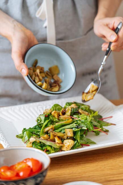 Crop female with bowl and spoon in hands adding fried squash into plate with green leaves while preparing salad at wooden table — Stock Photo