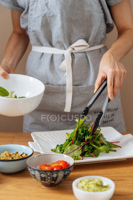 Erntehelferin mit Küchenzange legt frische grüne Blätter auf weißen Teller, während sie am Holztisch mit Zutaten steht und Salat zubereitet — Stockfoto