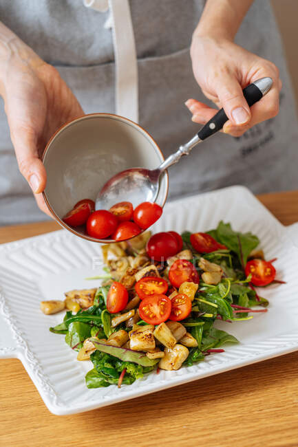 Crop female preparing healthy vegetarian salad and putting cut ripe tomatoes cherry on white plate with ingredients — Stock Photo