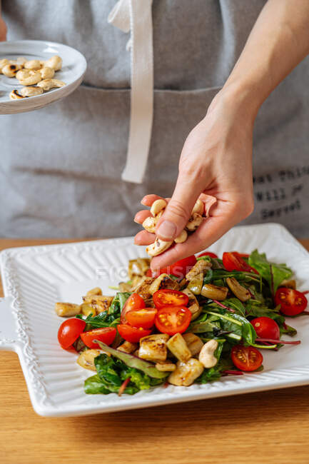 Crop female finishing delicious healthy colorful vegetarian salad and garnishing dish with roasted cashew nuts — Stock Photo