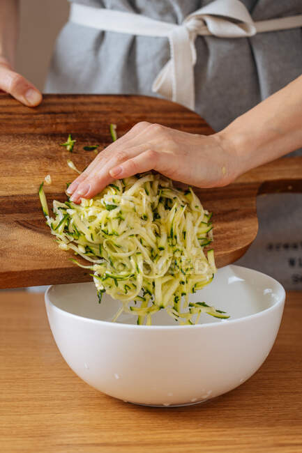 Crop casalinga mettendo zucchine griglia dal tagliere di legno in grande ciotola bianca durante la preparazione di cibo un tavolo di legno — Foto stock