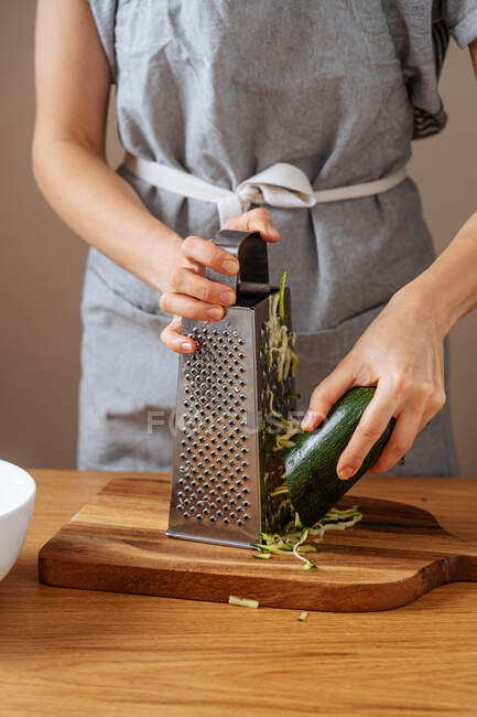Crop female in grey apron grating fresh zucchini while standing at wooden table in kitchen and preparing healthy dinner — Stock Photo