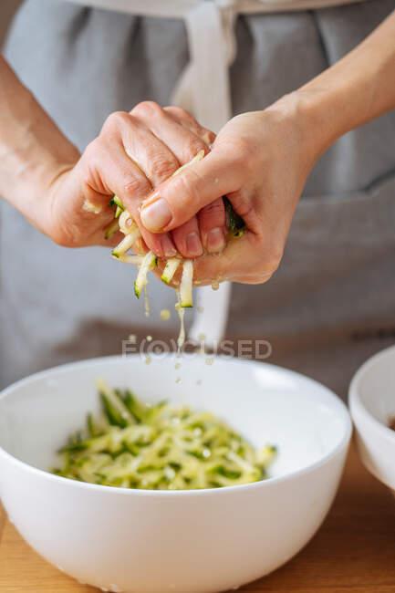 Crop hands of female squeezing fresh grating zucchini over white bowl while preparing food in kitchen — Stock Photo