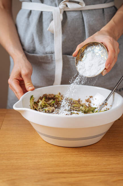 Cultivar hembra añadiendo sal en un tazón blanco con ingredientes mezclados mientras se prepara la cena en la mesa de madera en la cocina - foto de stock