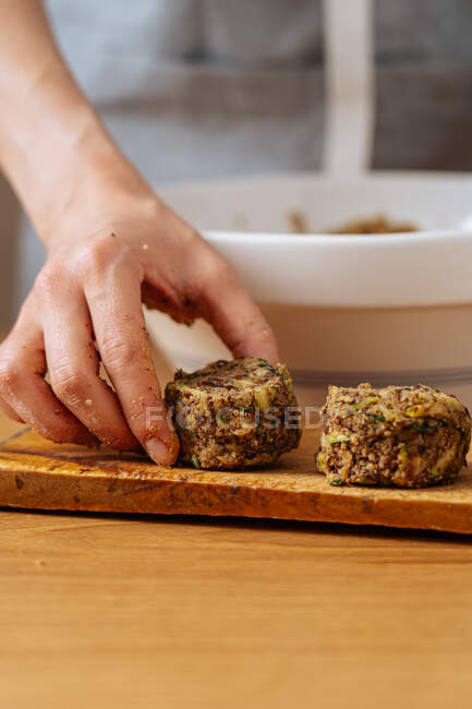 Crop hand of female putting raw vegan lentil and zucchini burger on wooden board tout en préparant le dîner à la table en bois — Photo de stock