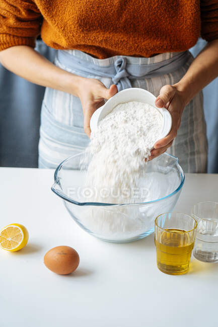 Crop housewife putting white flour into large glass bowl while preparing dough at white table with prepared ingredients — Stock Photo